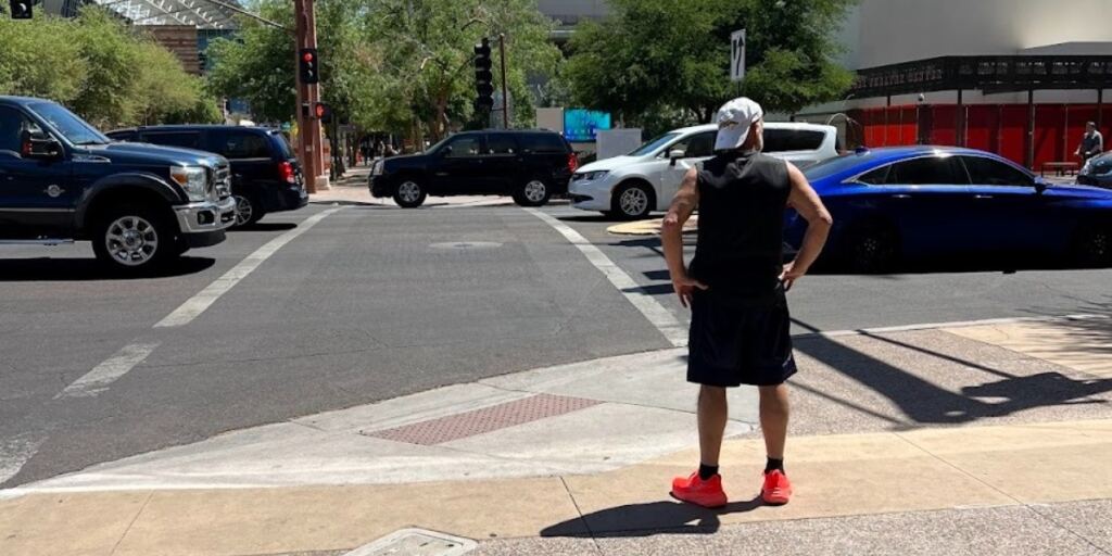 This image captures a person standing at an urban crosswalk on a sunny day, with a cityscape including various buildings and a Bank of America tower in the background. The scene highlights the urban environment and infrastructure, emphasizing pedestrian activity and city life.