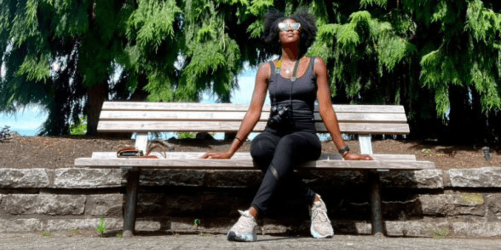 Whitney Mayfield sitting on a park bench, surrounded by tall trees, featured on the Urbanism by Chance podcast.